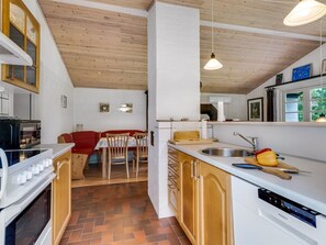 A kitchen with a wooden ceiling, white walls, and a herringbone tile floor. There is a dining area with red chairs and a table, and a kitchen counter with a sink and a dish rack. - "Söffren" - 450m to the fjord by Interhome (Øster Assels)