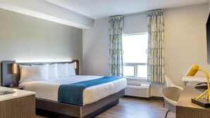 Une chambre d’hôtel moderne équipée d’un lit, d’un bureau, d’une chaise et d’une grande fenêtre agrémentée de rideaux à motifs.