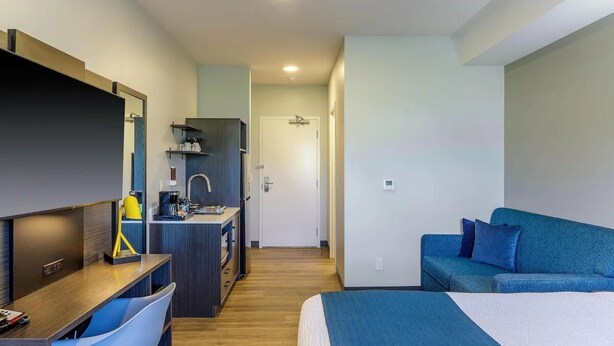 A hotel room with a bed, a desk with a chair, a kitchenette, and a blue sofa. - King Room with Kitchen | Free Parking | Quick 0.5-Mile Drive to Cedar Point (Hornepayne)
