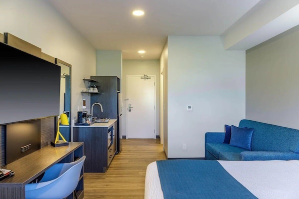 A hotel room with a bed, a desk with a chair, a kitchenette, and a blue sofa. - King Room with Kitchen | Free Parking | Quick 0.5-Mile Drive to Cedar Point (Hornepayne)