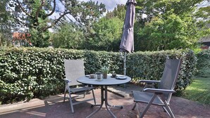 Een terras buiten met een ronde tafel en twee stoelen, omringd door een haag en bomen.