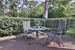 An outdoor patio with a round table and two chairs, surrounded by a hedge and trees. - Vrbo Property (Langeoog)