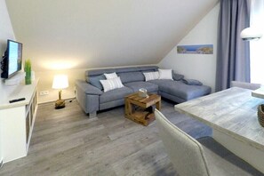 Ein modernes Wohnzimmer mit einem grauen Ecksofa, einem Couchtisch aus Holz und einem Fernseher auf einem weißen Lowboard.