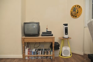 A room with a wooden table holding a television, a DVD player, and a bookshelf filled with DVDs. A white portable heater is on a small table next to the heater. - London Cozy Apartment Retreat (London)