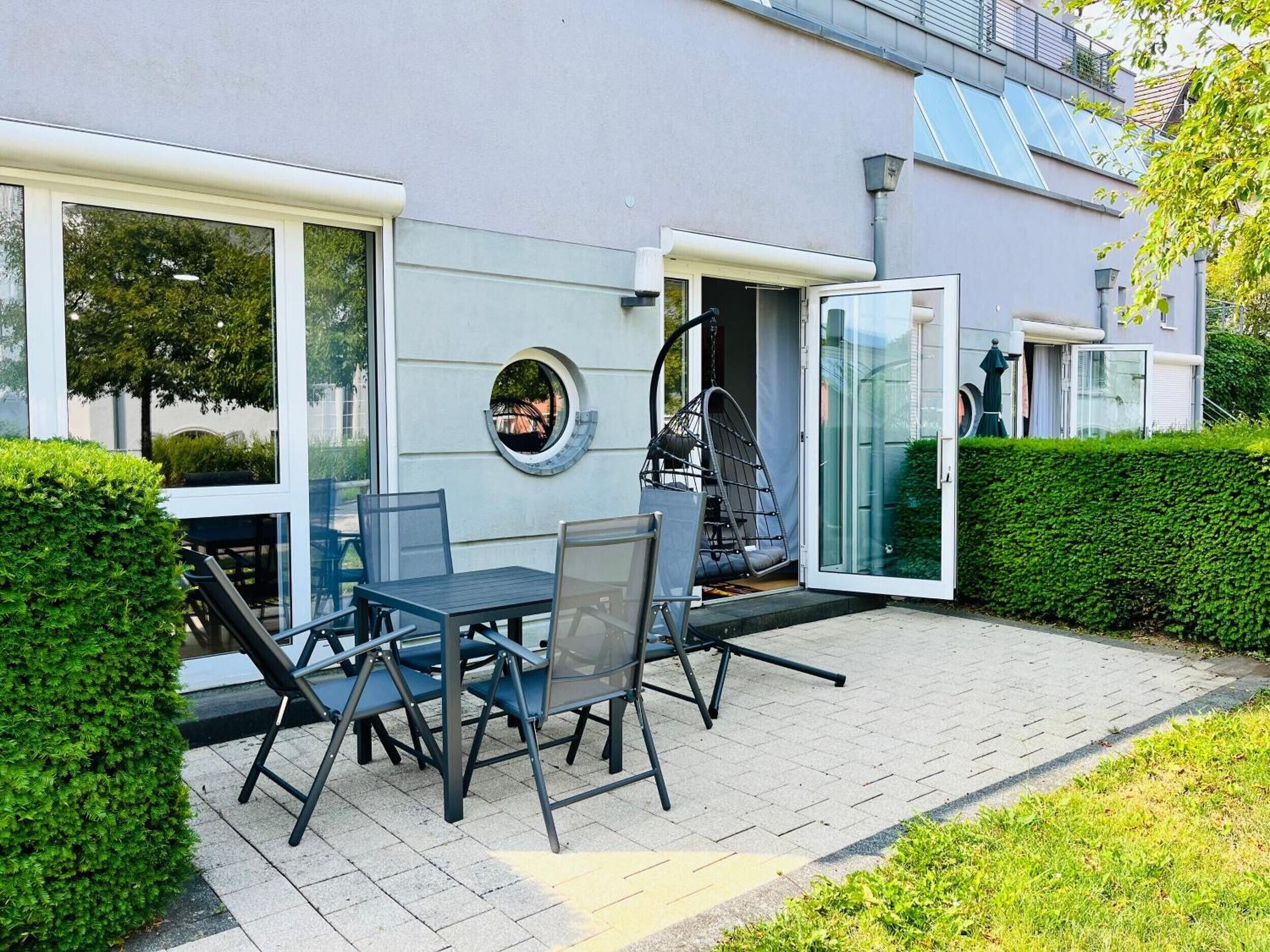 Eine moderne Terrasse im Freien mit Tisch und Stühlen, einem Hängesessel und einer Glastür, die zu einem überdachten Bereich führt.