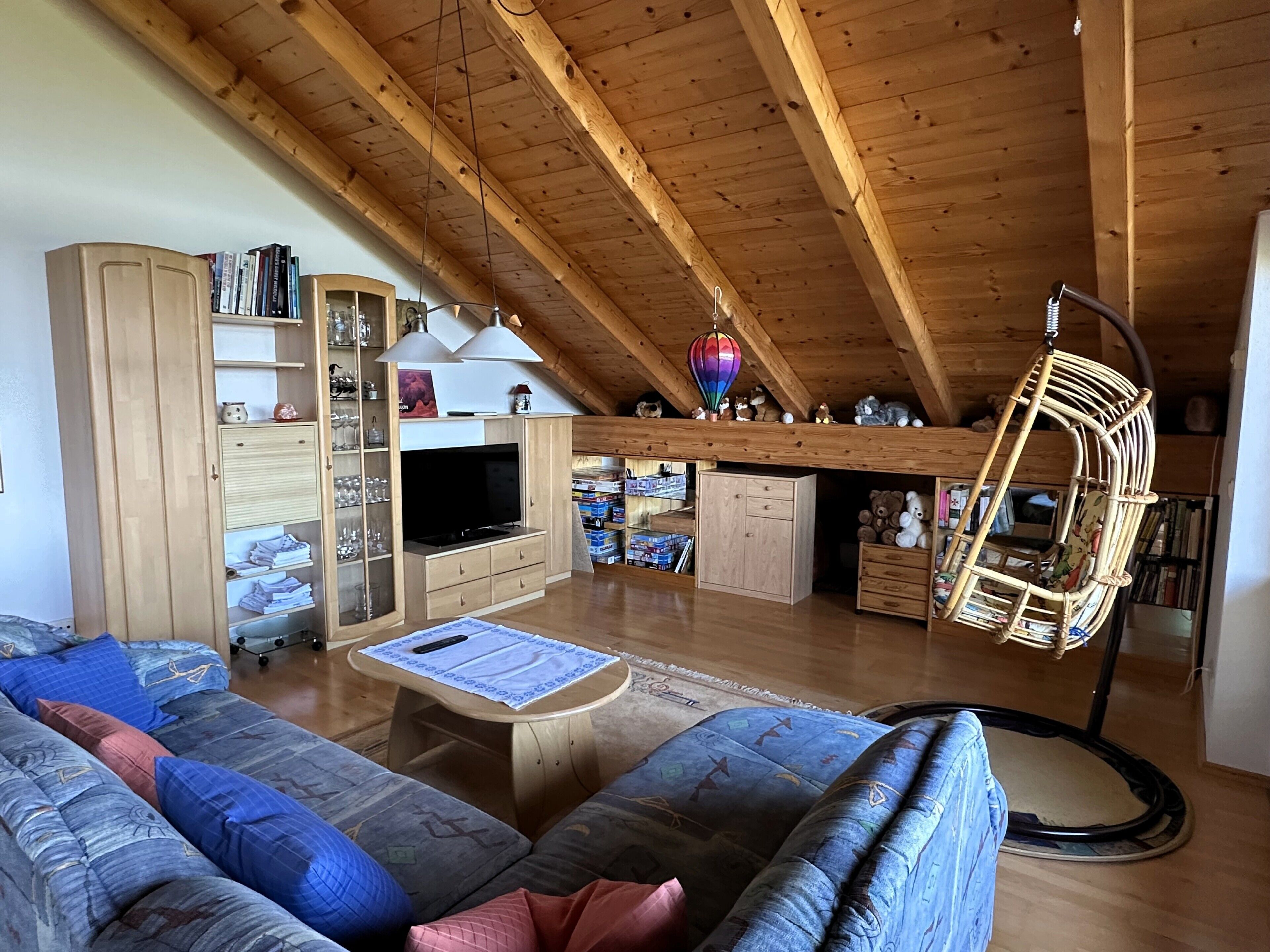 Ein gemütliches Wohnzimmer im Loft-Stil mit hohen Decken, Holzbalken, einer Couch, einem Fernseher und einem Hängesessel.