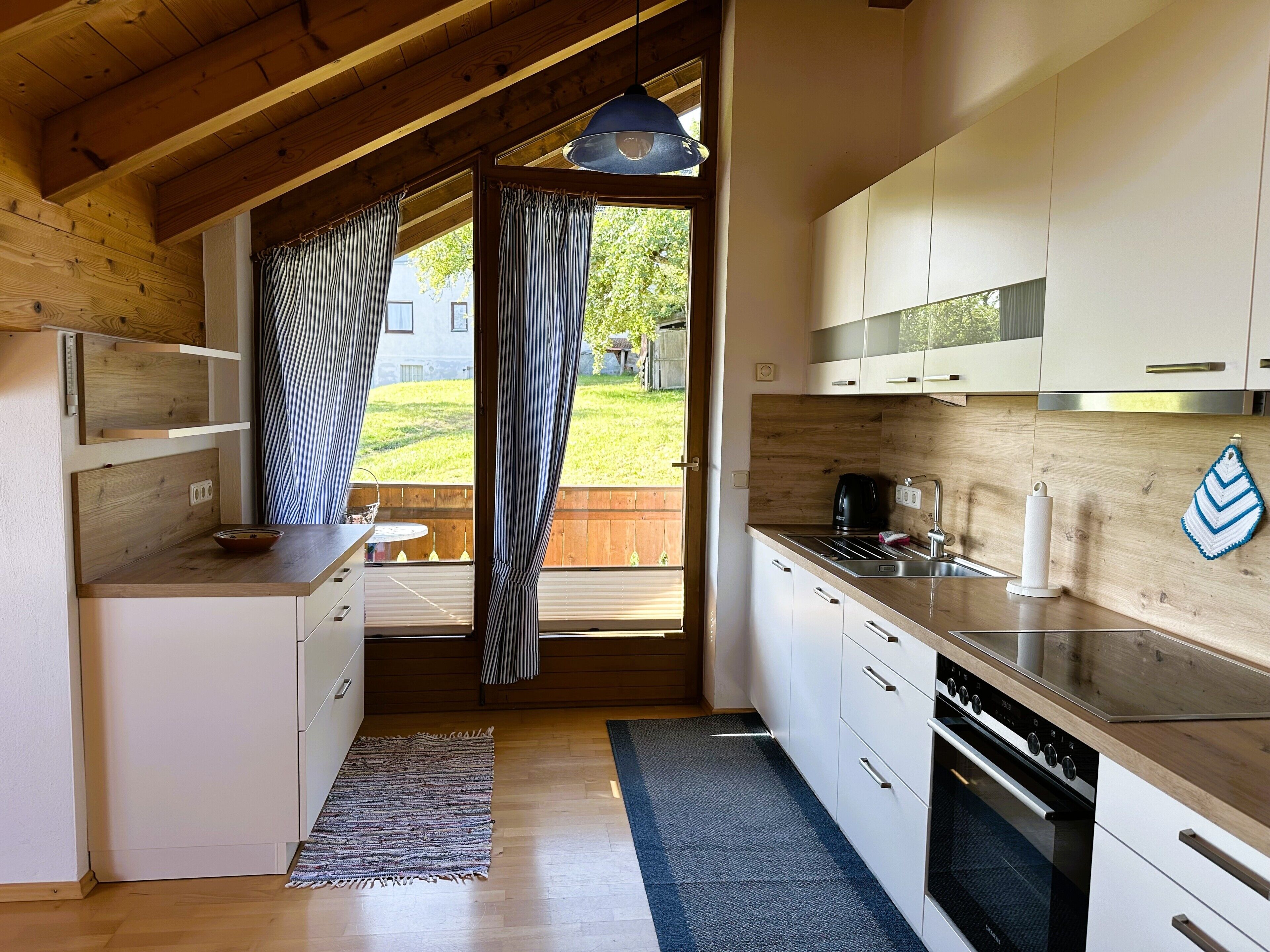Una cocina moderna con vigas de madera, un ventanal con cortinas, horno empotrado y armarios blancos.