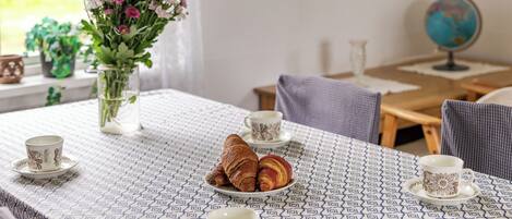 A table with a patterned tablecloth, two cups, a plate with croissants, and a vase with flowers.