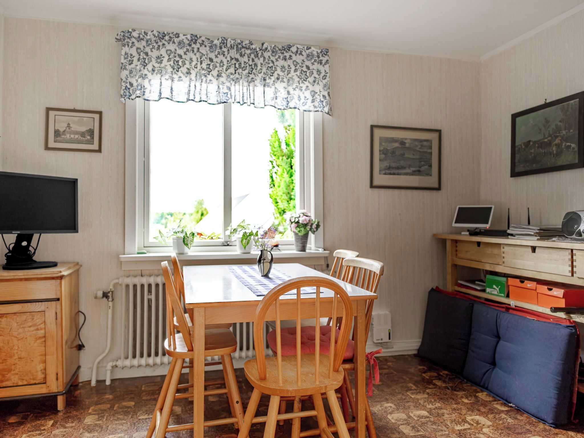 Ein Essbereich mit Tisch und Stühlen, einem Heizkörper, einem Fenster mit Blumenverhang und einem Schreibtisch mit einem Laptop.