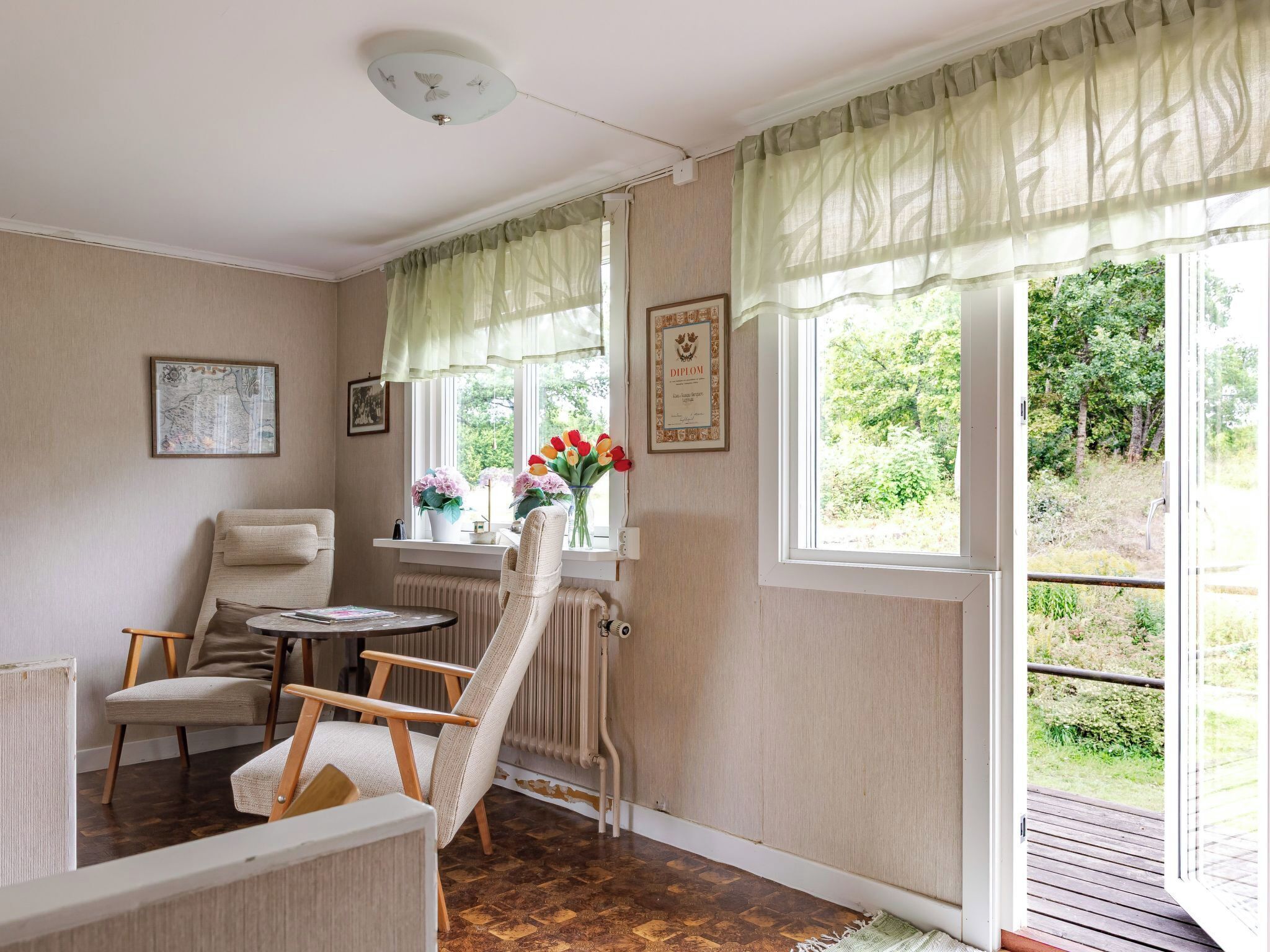A cozy room with a chair, a small table, and a radiator. There are two windows with curtains, and a framed diploma on the wall.