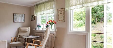 A cozy room with a chair, a small table, and a radiator. There are two windows with curtains, and a framed diploma on the wall.