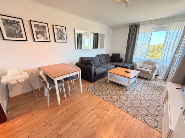 Ein Wohnzimmer mit einem Holzesstisch, einer Couch, einem weißen Sessel und einem gemusterten Teppich.