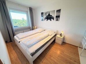 Ein Doppelbett mit Kopfteil, ein Nachttisch mit Lampe und ein Fenster mit Vorhängen.