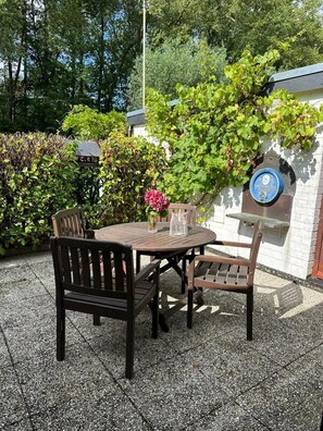 Un patio esterno con un tavolo rotondo in legno e quattro sedie, circondato da un pavimento in ghiaia e un siepe.