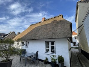 Una casa bianca con tetto di paglia, un piccolo patio con tavolo e sedie, e una stretta via.