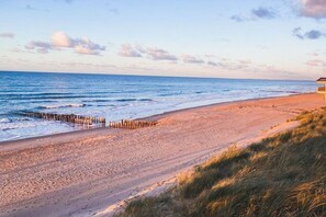 Ein Strand mit einer Holzsteganlage, sandigem Ufer und einem Gebäude in der Ferne.