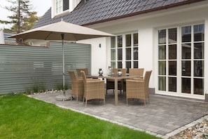 En udendørs terrasse med et bord, stole og en stor parasol, samt et hus med hvide facader og et gråt tag.