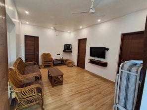 A living room with a sofa set, a coffee table, a television mounted on the wall, and a ceiling fan. - NT Grand Inn (Yercaud)