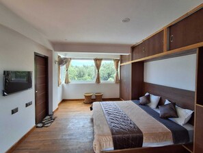 Un dormitorio con una cama, televisor, ventana con cortinas y piso de madera.