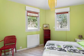 A bedroom with two windows, a radiator, a red chair, a bedside table with a lamp, and a bed with a patterned bedspread. - Holiday Home 'Maison Kerjean' with Shared Terrace, Private Garden and Wi-Fi (Plouha)