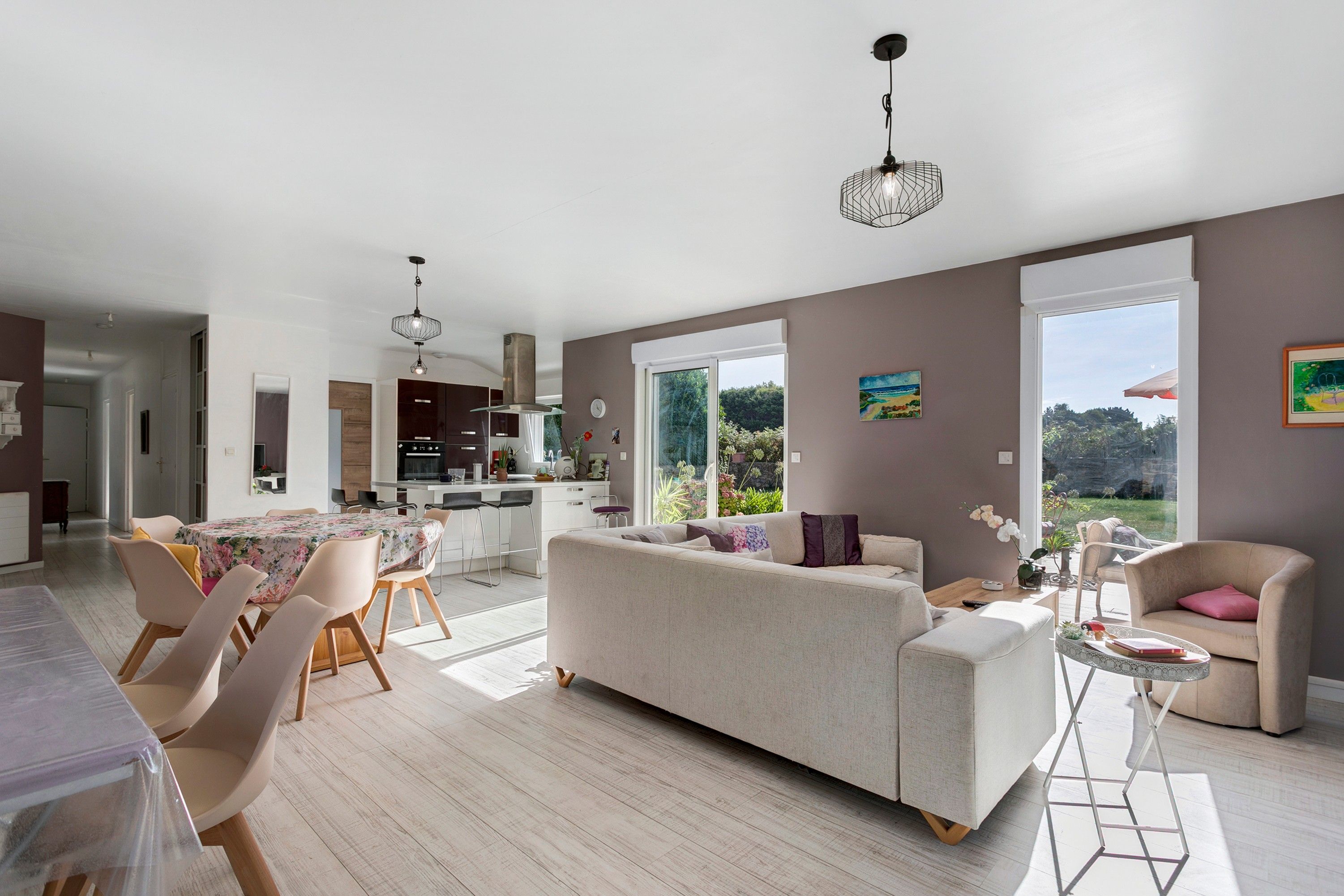 A spacious living area with a sofa, dining table, and chairs, a kitchen in the background, and large windows offering views of the outdoors.
