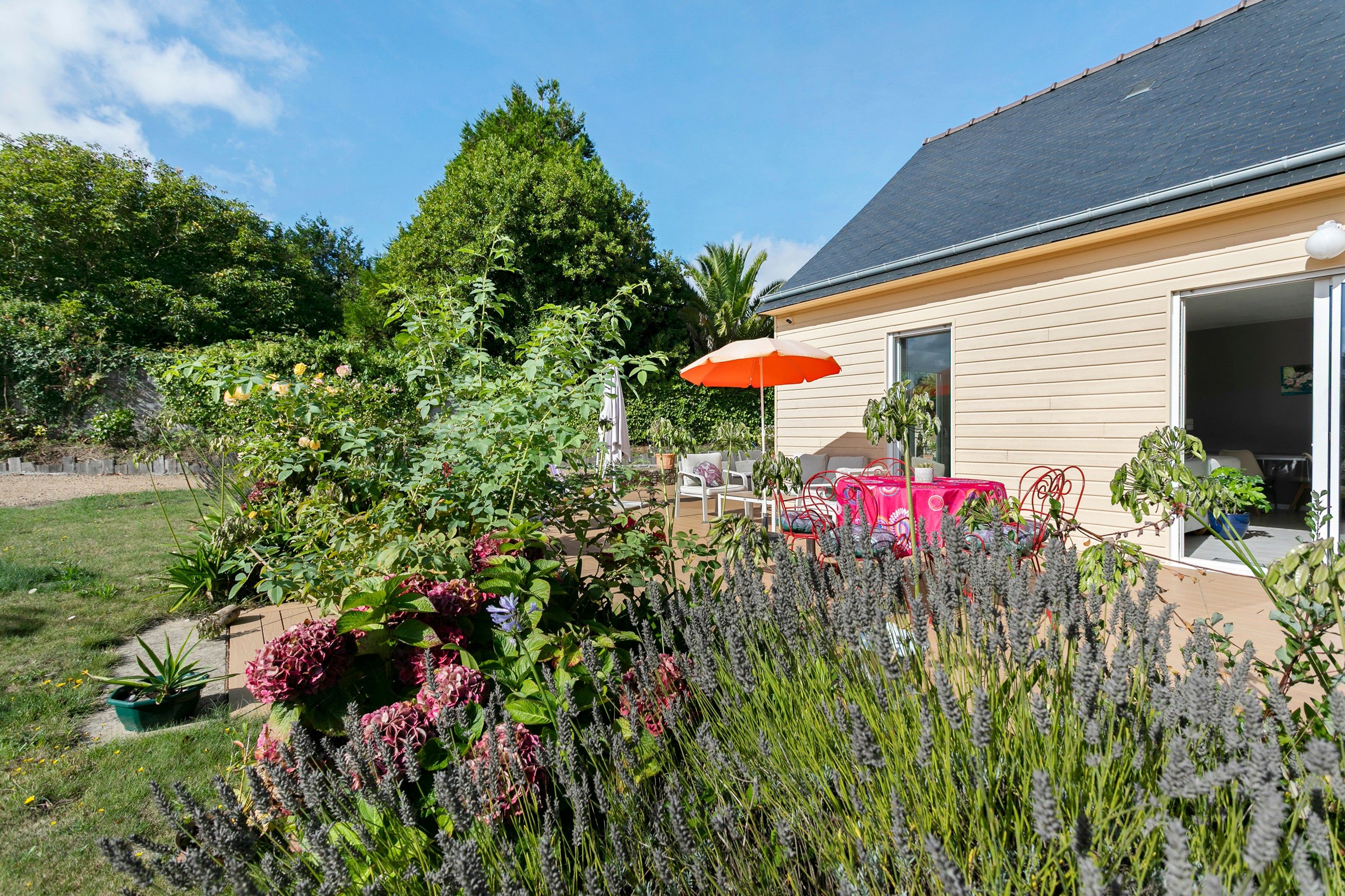 A house with a garden featuring various plants and flowers, a patio with outdoor furniture, and a red umbrella.