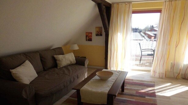 En stue med sofa, et bord med skål, en lampe og udsigt gennem et vindue med gardiner.