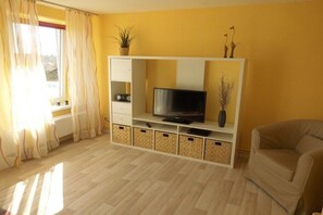 Ein Wohnzimmer mit einem Fernseher auf einer weißen Unterhaltungseinheit, einem beigen Sofa und einem Fenster mit Vorhängen.