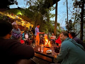 Un área de comedor al aire libre con una fogata y mesas, rodeada de árboles y un edificio con luces.