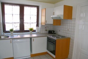 A compact kitchen with a dishwasher, oven, and a toaster. There is a window with a curtain and two potted plants on the counter. - The Comfortable Holiday Apartment (Kappeln)