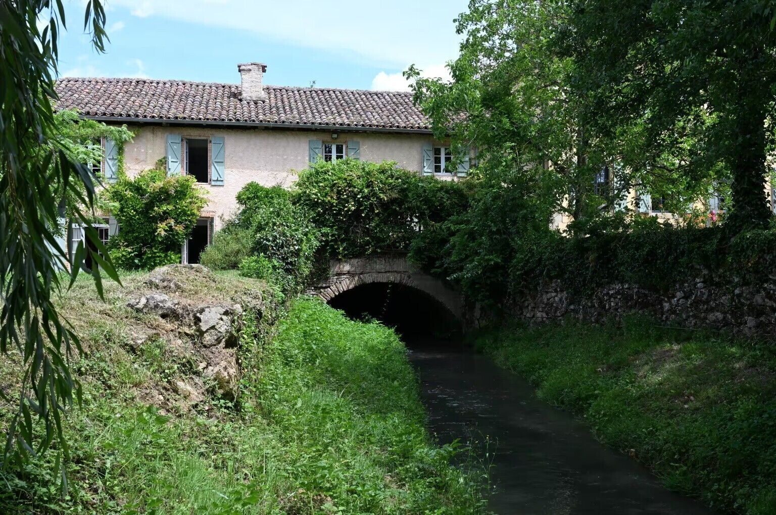 A rustic stone bridge over a narrow waterway, surrounded by lush greenery and a traditional building with a tiled roof.