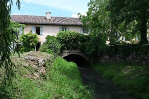 Un pont de pierre rustique enjambe un cours d’eau étroit, et il est entouré d’une végétation luxuriante et d’un bâtiment traditionnel doté d’un toit en tuiles.