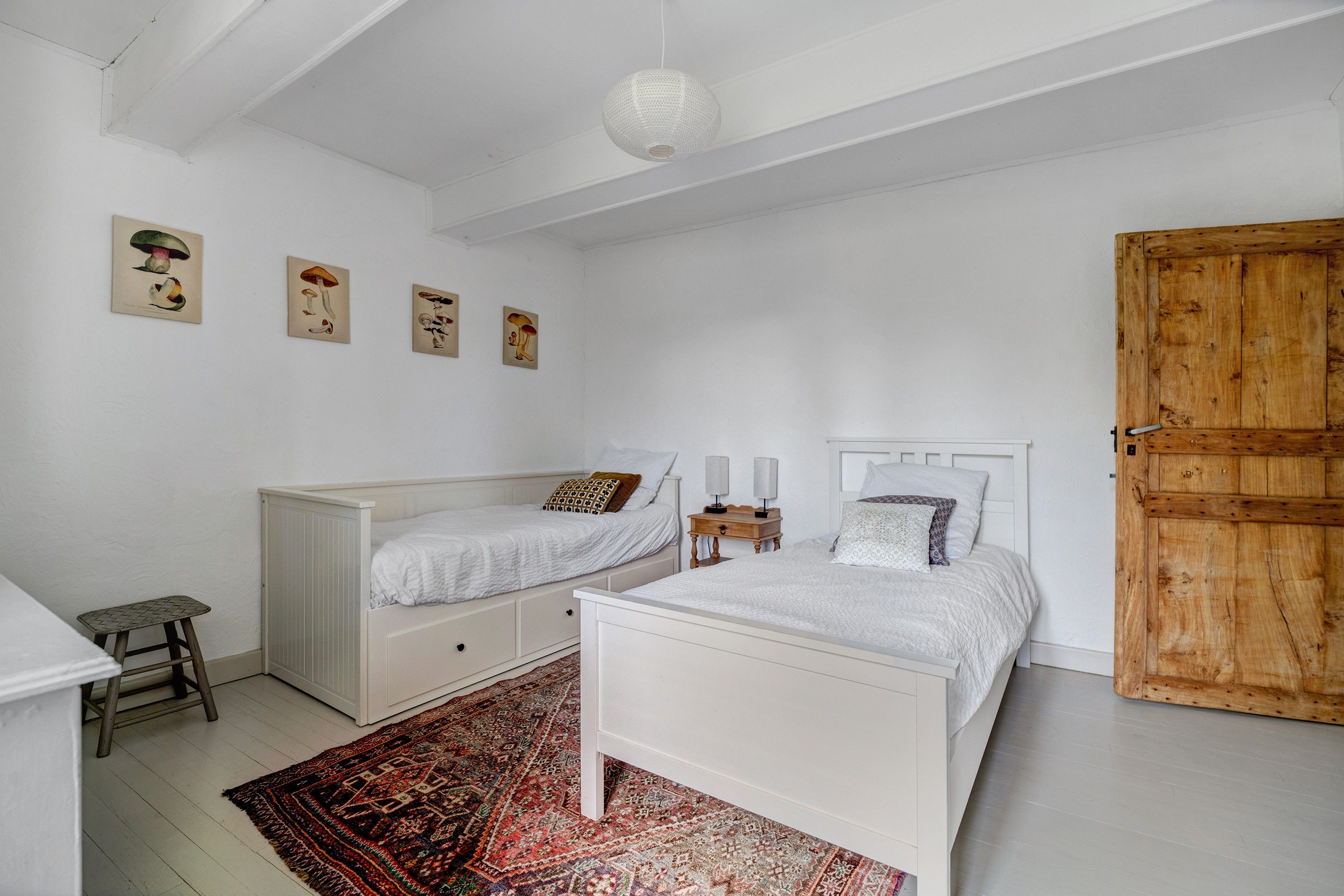 Een slaapkamer met twee eenpersoonsbedden, een houten garderobe, een klein krukje en een kamerkleed met dessin.