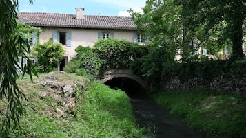 Uma ponte de pedra rústica sobre um pequeno curso d'água, rodeada por vegetação exuberante e um edifício tradicional com telhado de azulejos.
