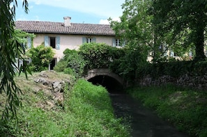 A rustic stone bridge over a narrow waterway, surrounded by lush greenery and a traditional building with a tiled roof.