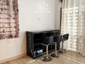 A room with a black entertainment unit, three black bar stools, a wall-mounted logo reading 'NATURE HOME', and a window with sheer curtains. - Casa 20 by Nature Home (Kuantan)
