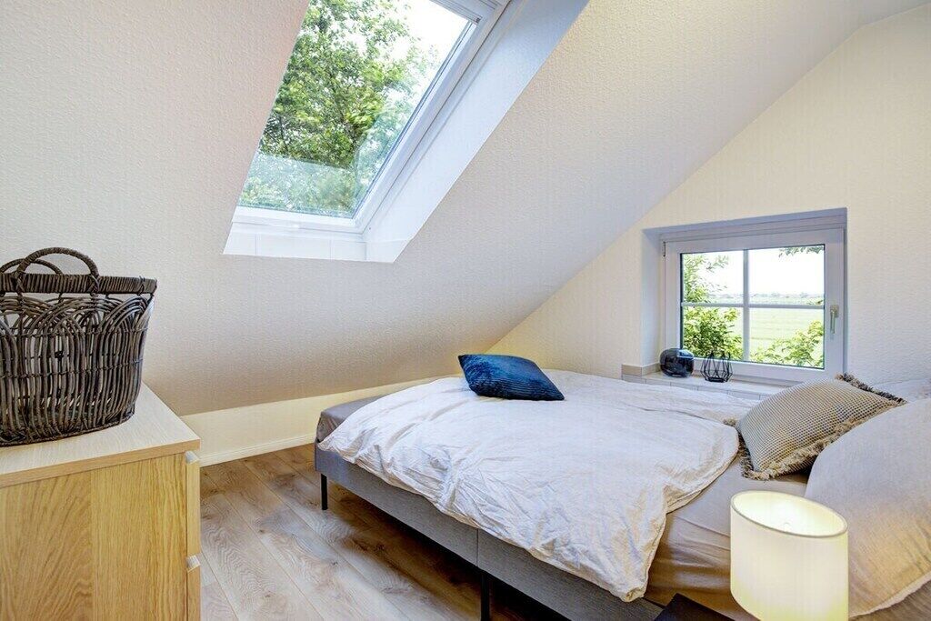 A bedroom with a sloped ceiling, a bed with white bedding, a bedside table with a lamp, and a window with a view of greenery.