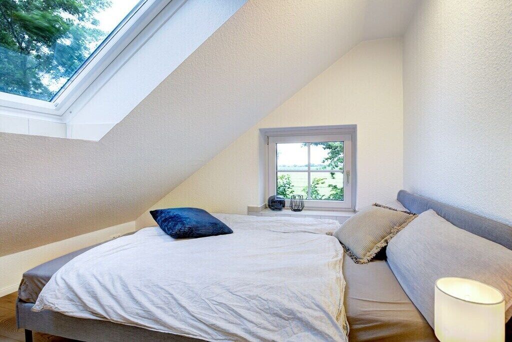 A bedroom with a sloped ceiling, a window, a bed with pillows, and a bedside lamp.