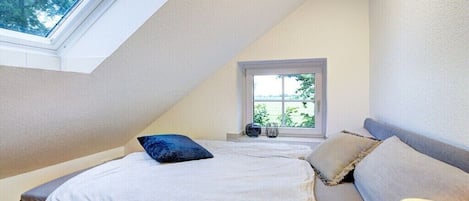Un dormitorio con techo inclinado, una ventana, una cama con almohadas y una lámpara de noche.