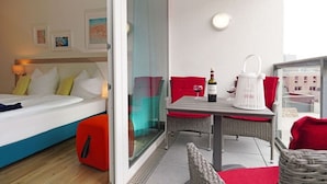 Habitación de hotel con una cama, una mesa con una botella y un vaso, y un balcón con muebles de mimbre.