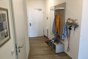 Ein Flur mit einer weißen Tür, einer Bank, einem Kleiderständer mit Kleidung und einem an der Wand befestigten Regal mit Gegenständen.