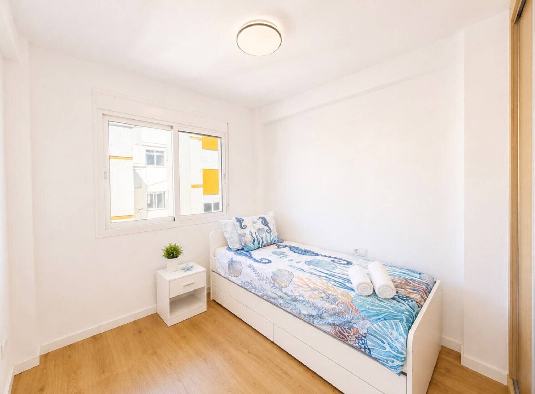 Una habitación pequeña con cama individual, suelo de madera, mesita de noche blanca con una planta, ventana con persianas amarillas y una lámpara en el techo.