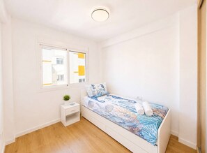Une petite chambre avec un lit simple, un plancher en bois, une table de chevet blanche ornée d’une plante, une fenêtre avec des stores jaunes et un luminaire au plafond.
