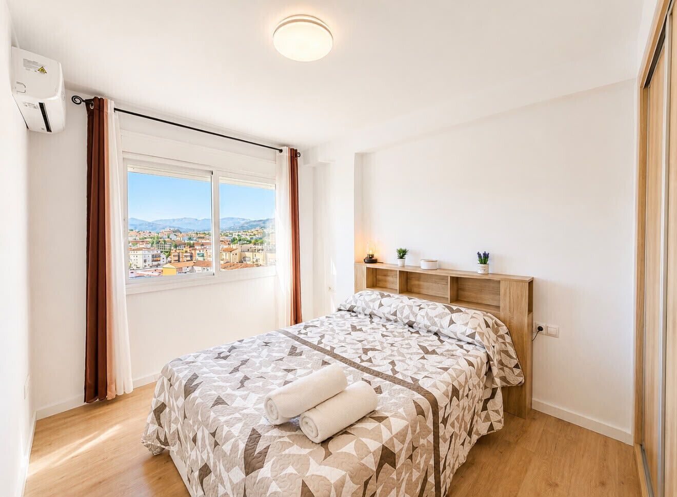 A bedroom with a bed, a window with curtains, a bedside table with a lamp, and a view of a cityscape.