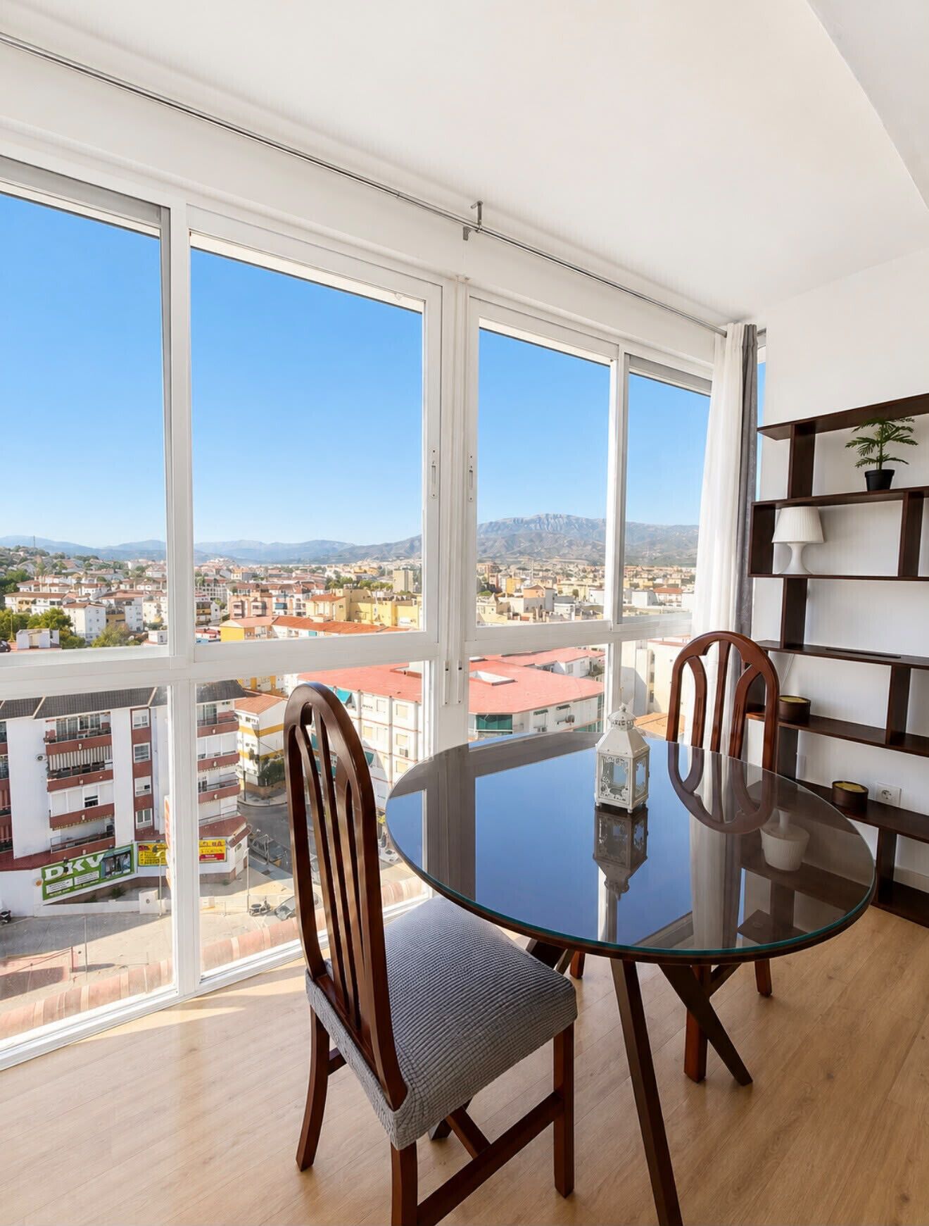 A dining area with a glass table and chairs, large windows with a city view, and a bookshelf.