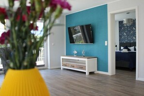 A modern living room with a teal accent wall, a flat-screen TV mounted above a white console, and a bed with a dark blue bedspread in the background. - Bernstein Suite & Breakfast Apartment 04 (Büum)