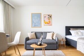 Una habitación de hotel moderna con un sofá gris, una mesita redonda pequeña, una cama con cabecero oscuro y dos obras de arte en la pared.
