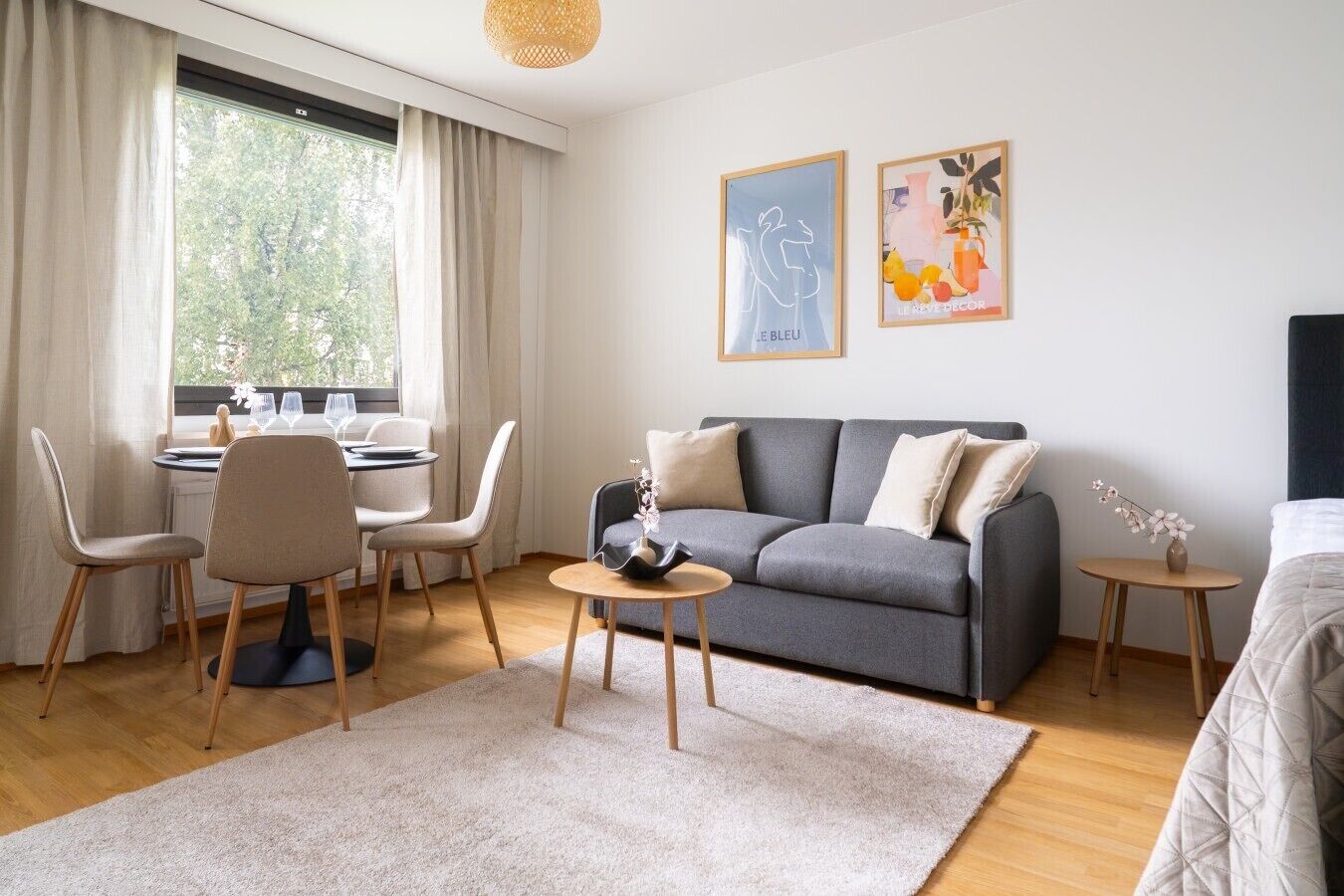 A modern hotel room with a grey sofa, a dining table with chairs, a small side table, and a bed.