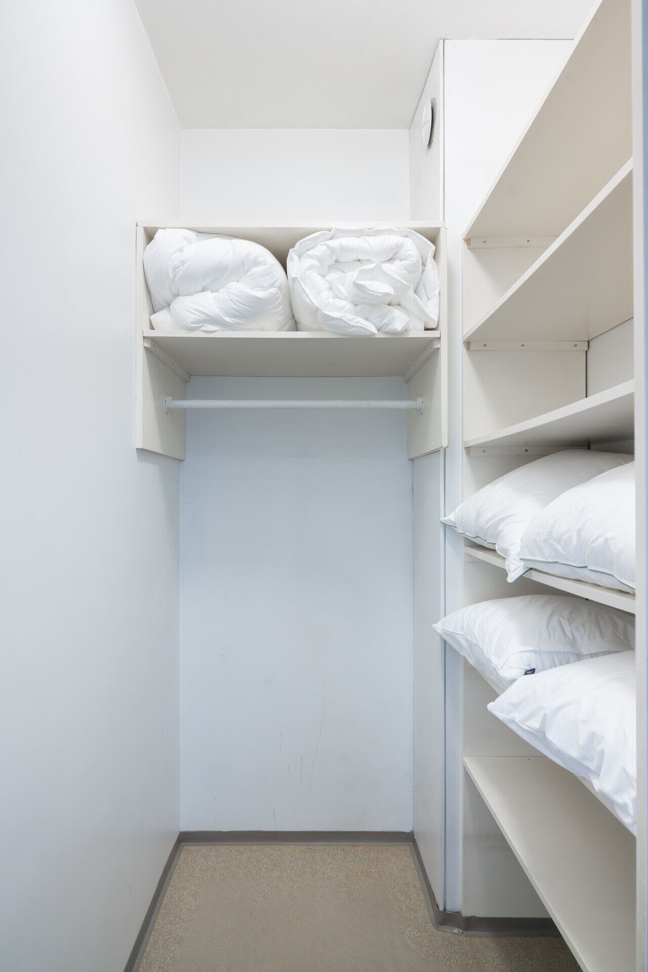 A small, white closet with shelves and a hanging rod, containing neatly folded white bedding.
