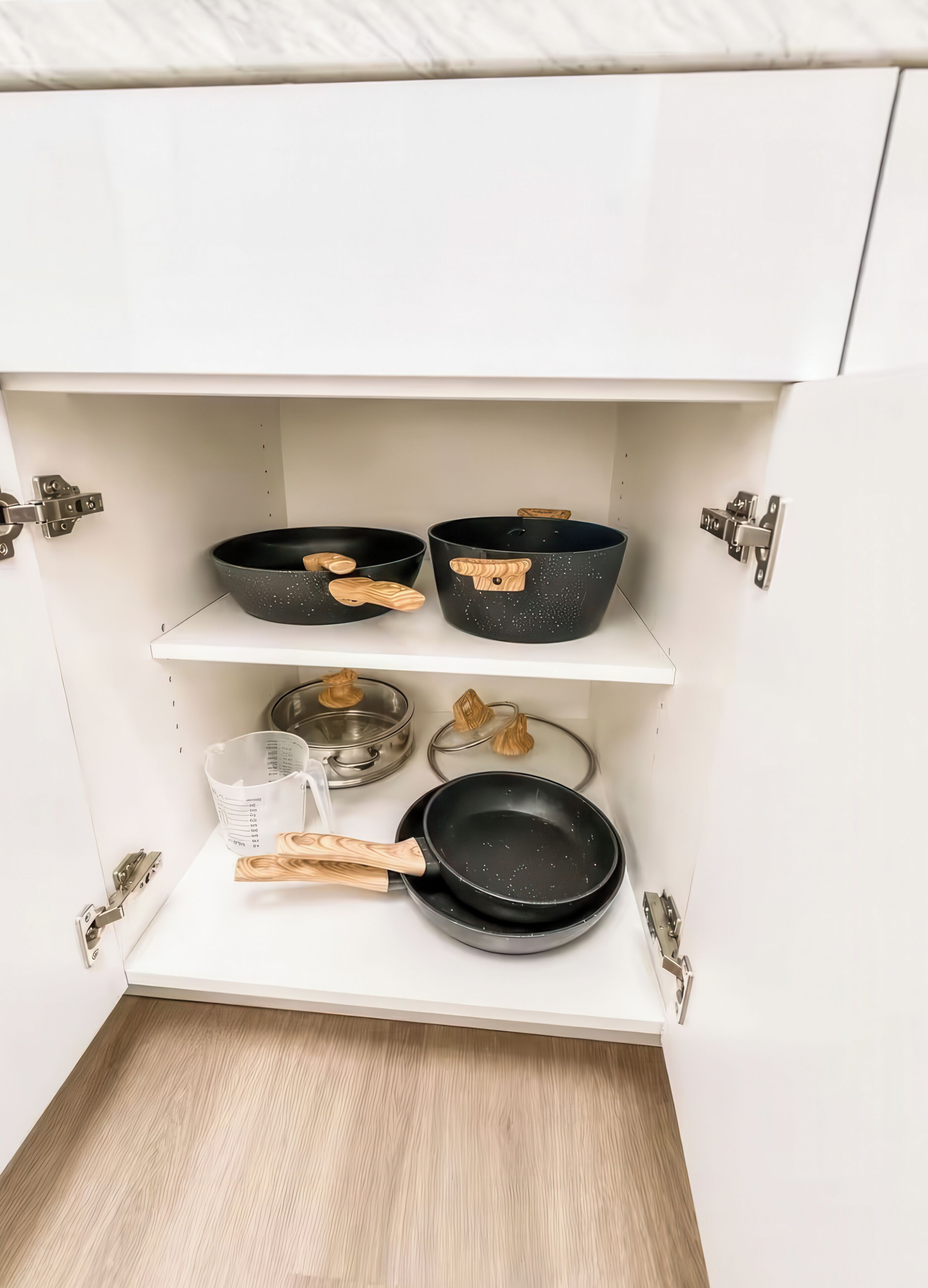 A kitchen cabinet with three black frying pans and a measuring cup.